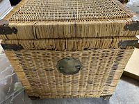 Close-up of front of trunk showing woven wicker texture and oval brass latch plate with no lock.