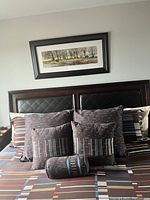 Framed landscape print mounted on bedroom wall above bed with brown padded headboard and pillows