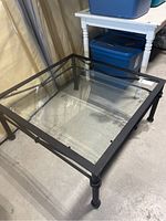 Square metal frame coffee table with clear glass top shown from side angle on a concrete floor.