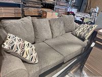 Front angle view of gray upholstered three-seat couch with rolled arms and two patterned throw pillows sitting on the seat cushions.
