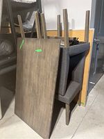 Grey wooden table top leaning against stacked grey upholstered chairs, showing table surface and chair legs with screws for assembly.