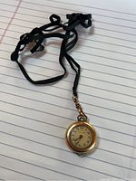 Full view of ladies gold-filled round pocket watch on a black fabric strap lying on lined paper.