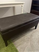 Full side view of dark brown leather-like rectangular storage bench with cushioned top and four legs, placed on concrete floor next to white table and wooden shelf.