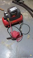 Photo showing the full view of the red and black King Canada air compressor sitting on a basement floor, with coiled red air hose and external black hose visible.