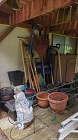 Photo showing large plastic rakes and shovels in various sizes, metal pick axe, plastic and metal gardening tools leaning against wall with planters and other supplies below.