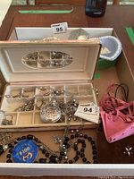 Open jewelry box showing several silver-tone necklaces, a silver-tone pocket watch on chain, black beaded necklace, pink suede bracelets, and a blue $100 casino chip. The box has an interior mirror.