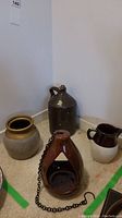 Four stoneware and ceramic items arranged on floor corner: brown jug, glazed pitcher, painted pot, hanging candleholder with chain.
