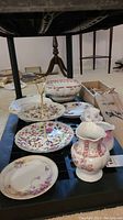 Wide angle photo showing assortment of serving dishes including two-tiered plate stand, pitchers, and plates on a black storage rack.