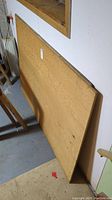 Plywood sheets leaning against a white wall in a basement, showing the smoother brown front side and visible wood grain.