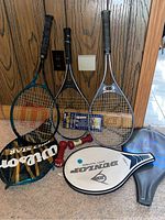 Photo of three tennis rackets and their cases, two red hand weights, and a racketball glove on a wooden floor with wood paneled wall backdrop.
