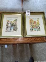 Pair of framed prints placed side by side on a wooden surface, showing both artwork and matching gold frames.