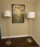 Two standing floor lamps against a beige wall with floral artwork behind. Both lamps off. One lamp has white shade with silver lines, other has plain white shade and glass detail on stand.