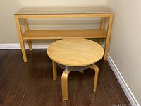 Rectangular wood table with glass top and wooden shelf beside small circular wood table with glass undershelf photographed on hardwood floor against beige wall.