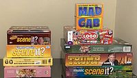 Photo of stacked board games showing Comedy Movies Scene It?, Puzzle box, and other games with titles visible like Mad Gab and Trump The Game.