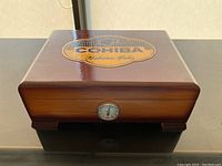 Closed wooden cigar storage box with polished finish and large Cohiba logo on top, analog humidity sensor on front center edge.