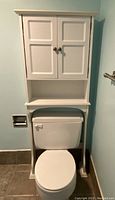 Front view of white over toilet storage cabinet fitted over a toilet showing closed doors and open shelf above the toilet tank.
