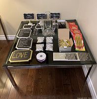 Photo showing an assortment of wedding decoration items on a glass table, including reusable chalkboard signs, clear vases filled with decorative crystals, rolls of ribbon, packages of cake sparklers, small wrapped chocolates, and decorative rhinestone bands.