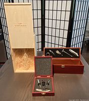 Photo showing two wooden wine boxes side by side, with foam inserts displaying multiple chrome and metallic wine tools including corkscrews, pourers, and stoppers. A branded cardboard sleeve is also included.