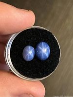 Two oval cabochon star sapphire gemstones with visible six-rayed star effect viewed on black felt backing inside small round container held in hand.