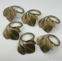 Group of six heavy brass napkin rings arranged on a flat surface, showing leaf design and solid brass construction.