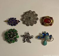 Photo showing all six vintage brooches on a beige background, various styles and colors including silver filigree, gold-tone with red stones, blue-purple cluster, green floral, silver swirl with rhinestones, and blue/turquoise with pearl (missing pin).