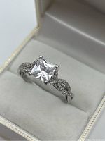 Front angle image of silver 925 ring with large clear princess cut stone in raised prong setting, placed inside white cushioned ring box.