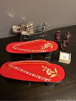 Photo showing a pair of red wooden sandals with floral designs, three small red Buddha figurines, a silver-coloured metal figurine of a man with a cart, and a small box with painted rice grains.