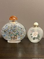 Photo showing two Chinese interior painted snuff bottles: the larger featuring colorful butterflies and flowers with a brown stopper, the smaller featuring a white flower and calligraphy with a white stopper.