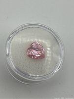 Pink heart shaped faceted topaz gemstone in clear container on white background.