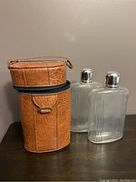 Closed brown textured leather zip-up case for the double flask, standing upright on a wooden surface with two clear glass flasks beside it.