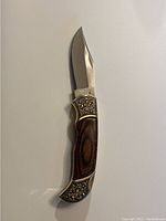Folding knife with wood handle held open showing the polished blade and engraved metal ends.