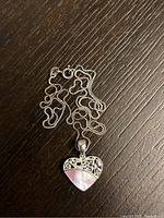 Sterling silver necklace with heart pendant laid out on a dark surface showing chain and clasp, heart pendant features pink mother of pearl and silver filigree design.