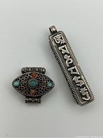 Front view of both sterling silver Tibetan prayer boxes; oval box with coral and turquoise inlay, rectangular pendant with script