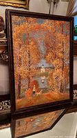 Frontal view of the framed painting showing orange and yellow autumn trees with a small white dome building reflected in a water body, woman and dog in foreground.