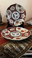 Side view of Imari porcelain plate on black display stand, showing floral and bamboo motifs in red, blue, and green with scalloped rim.