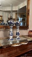 Pair of tall ironstone whale oil lamps with blue scenic designs, metal burner collars, and glass chimneys on a reflective antique table in front of a gold-framed mirror.