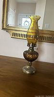 Amber glass oil lamp with scalloped chimney and metal base, photographed on a wooden surface with a mirror in the background.