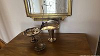 Three silver plated items on a wooden table including a bridal basket and two serving bowls.