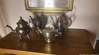 Photo of four antique silver teapots arranged on a wooden dresser beneath a framed mirror.