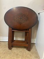 Photo of three folded oval wooden side tables with decorative black ornate pattern on tops stood vertically against the wall.