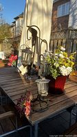 Two black metal candle holders with hanging glass lanterns seen on a patio table in sunlight, with a large white umbrella and flower pot in background.
