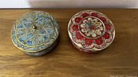 Two closed decorative tins with intricate floral and geometric patterns on lids, placed side by side on a wooden surface.