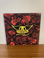 Front cover of Aerosmith Permanent Vacation vinyl album featuring black background with red illustrations and yellow Aerosmith logo and title.