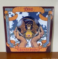 Front cover of Dio Sacred Heart vinyl album featuring a dragon in a crystal ball held by two hands, with orange ribbon banners and text.