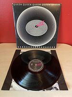 Front cover of Queen Jazz vinyl album with a white spiral graphic and pink arrow in the center on a black background. Vinyl record with label visible in front.