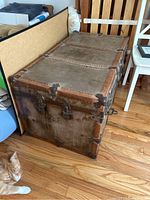 Side angle view of large antique wooden trunk with visible metal corner brackets and lock hardware, trunk closed, resting on wooden floor.