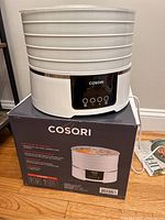 Cosori food dehydrator sitting on top of its box, showing front digital control panel with 4 buttons and 5 stackable white trays.