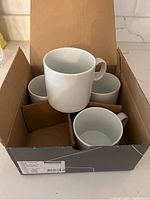 Four white ceramic mugs with handles in original partitioned box, product label visible indicating Thomas brand and Loft by Rosenthal Weiss line.