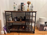 Front and side view of rolling bar cart with multiple shelves holding glass bottles, mugs, a brass vase with dried flowers, and a decorative cat sculpture.