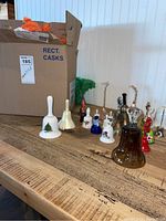 Photo of multiple bells in various styles and materials arranged on a wooden table with a cardboard box in the background